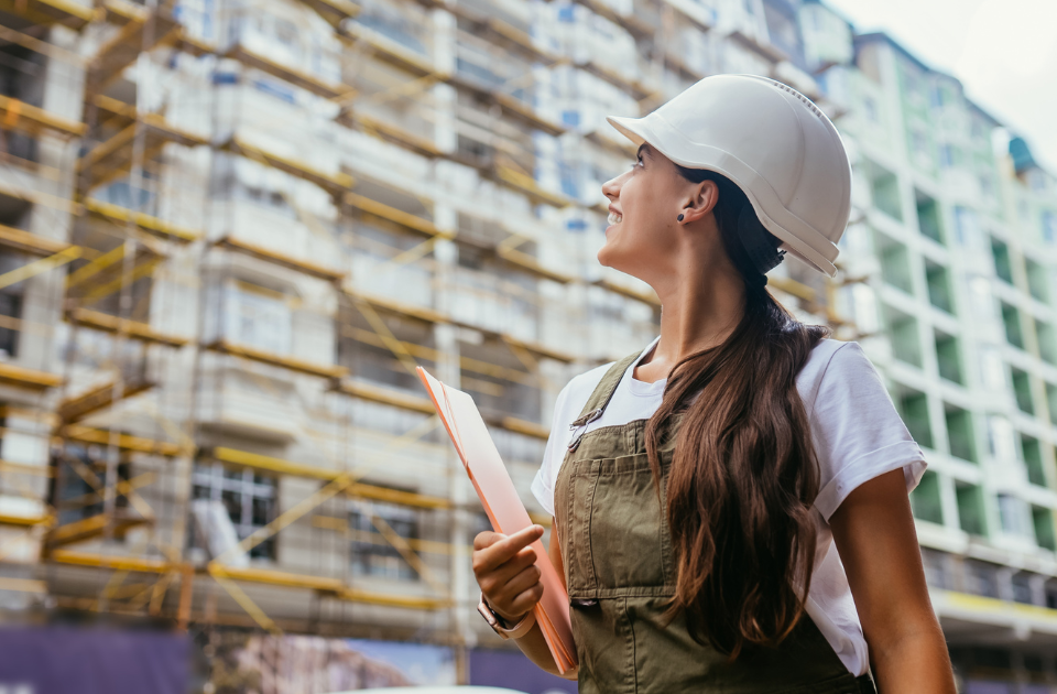 Uma mulher segurando uma prancheta, olhando para um prédio em construção. Ela está usando um capacete branco de obra.